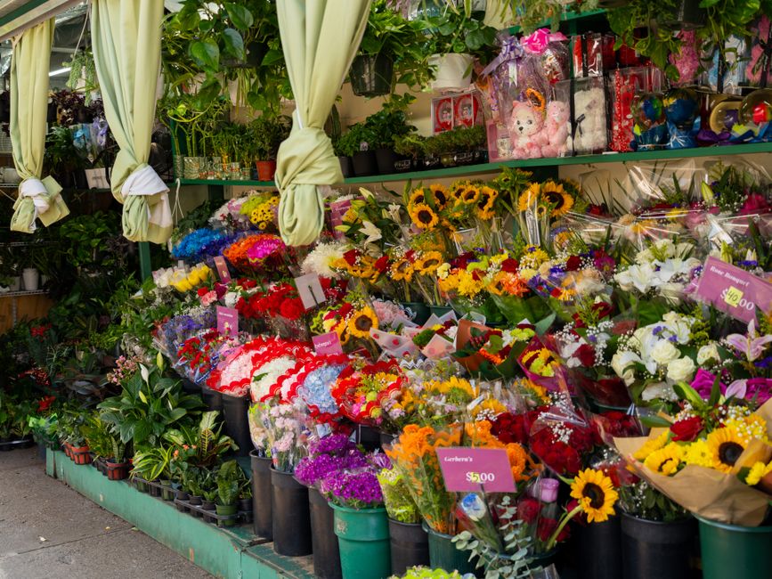 An outdoor floral display featuring a variety of fresh flowers arranged in bouquets and potted plants. The arrangement includes bright yellow sunflowers, mixed colorful blooms such as red roses, white