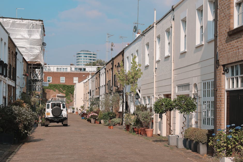 The image depicts a quiet residential cobblestone street lined with modern, white-painted townhouses on one side and partially building-covered structures on the other. Potted plants and small trees w