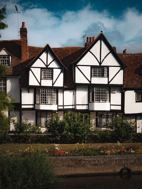 An exterior view of a traditional Tudor-style house with black and white timber framing and a dark tiled roof, set against a partly cloudy sky. The house features multiple small, sash-style windows. I