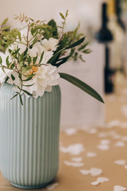 A close-up of a bouquet arrangement in a tall, ribbed, light green ceramic vase filled with white flowers, possibly freesias or gardenias, surrounded by green foliage and slender leaves. The arrangeme
