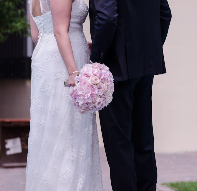 A close-up of a bride and groom during a wedding at Barking Park, featuring the bride in a white lace wedding dress holding a round bouquet of pink hydrangeas and roses. The bouquet is wrapped in soft